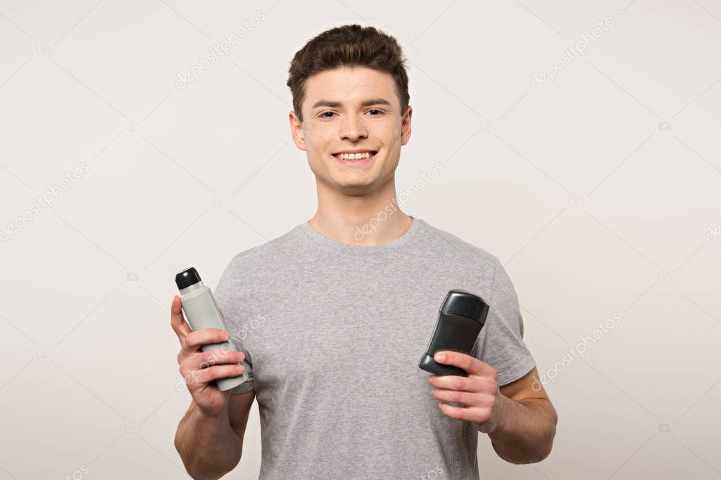 young man in grey t-shirt holding deodorants and smiling at camera isolated on grey