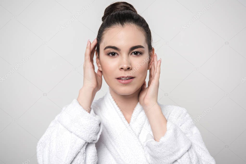 beautiful woman in bathrobe touching face while looking at camera isolated on grey