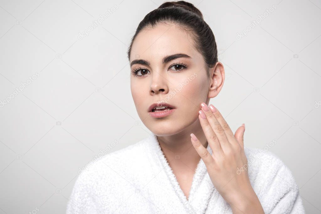 pensive woman in bathrobe touching face while looking at camera isolated on grey