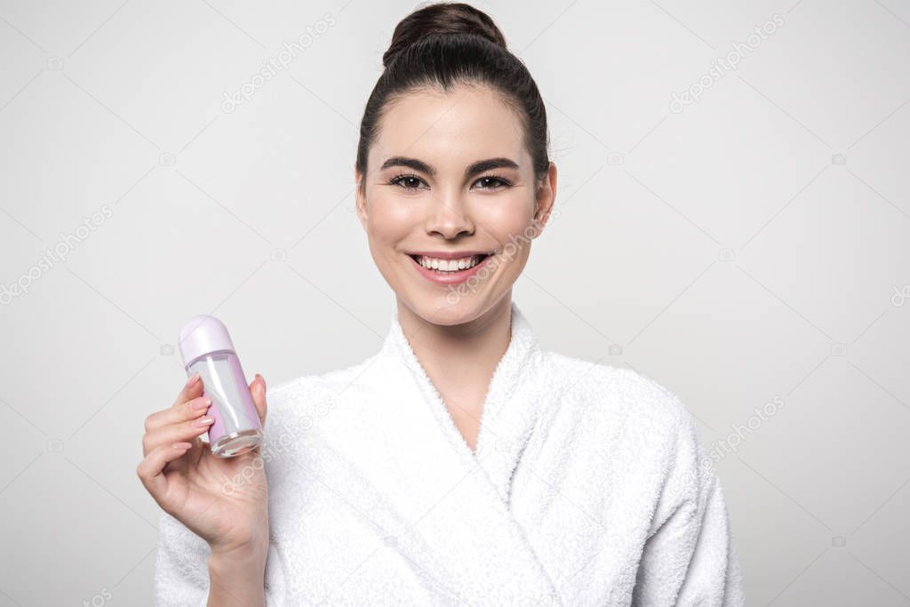 joyful woman in bathrobe smiling at camera while holding deodorant isolated on grey