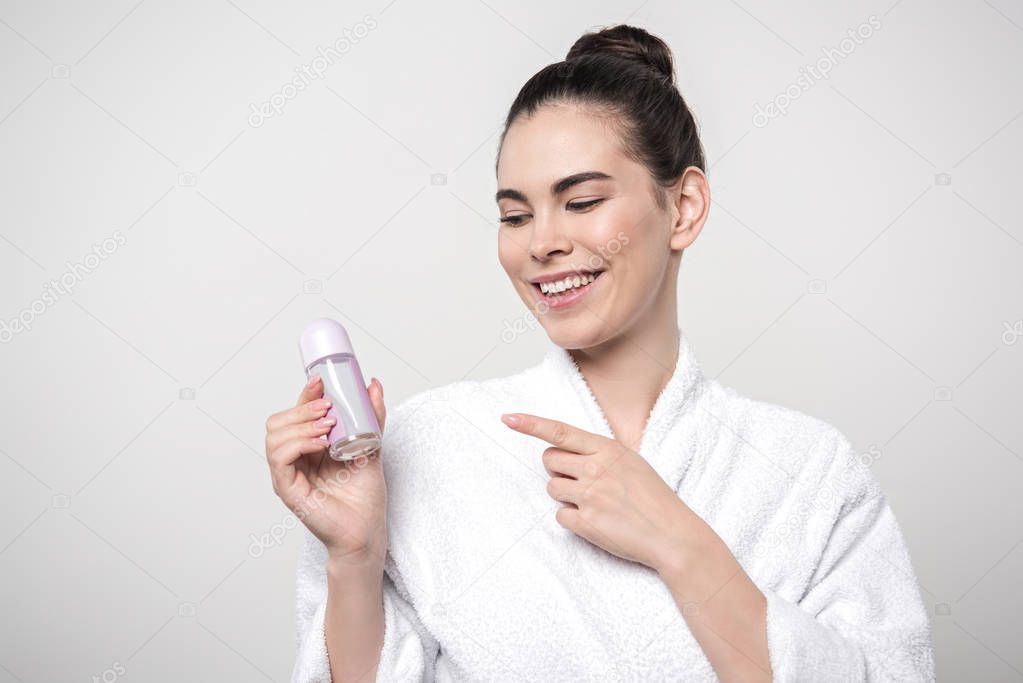 happy woman in bathrobe pointing with finger at deodorant isolated on grey