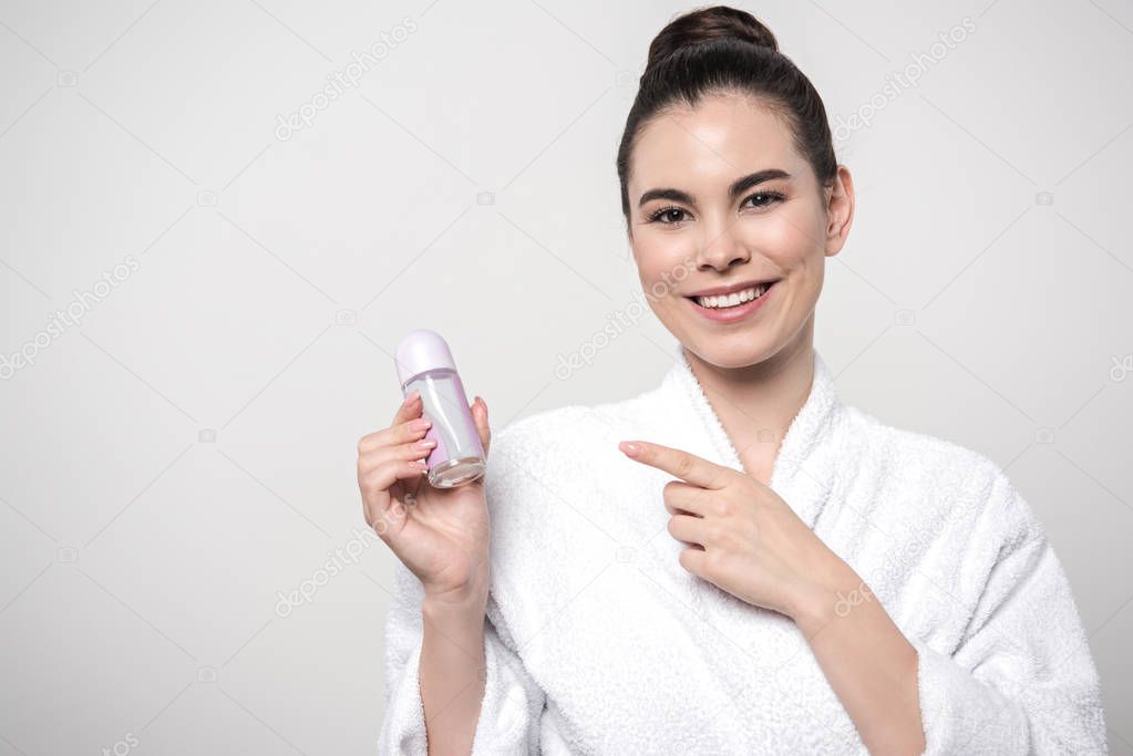 cheerful woman in bathrobe pointing with finger at deodorant while looking at camera isolated on grey