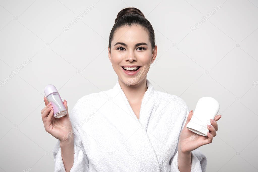cheerful woman in bathrobe holding deodorants while looking at camera isolated on grey