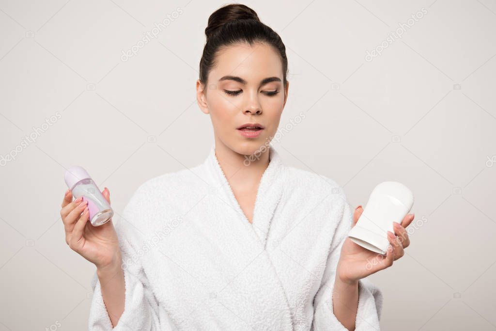 skeptical woman in bathrobe holding deodorants isolated on grey
