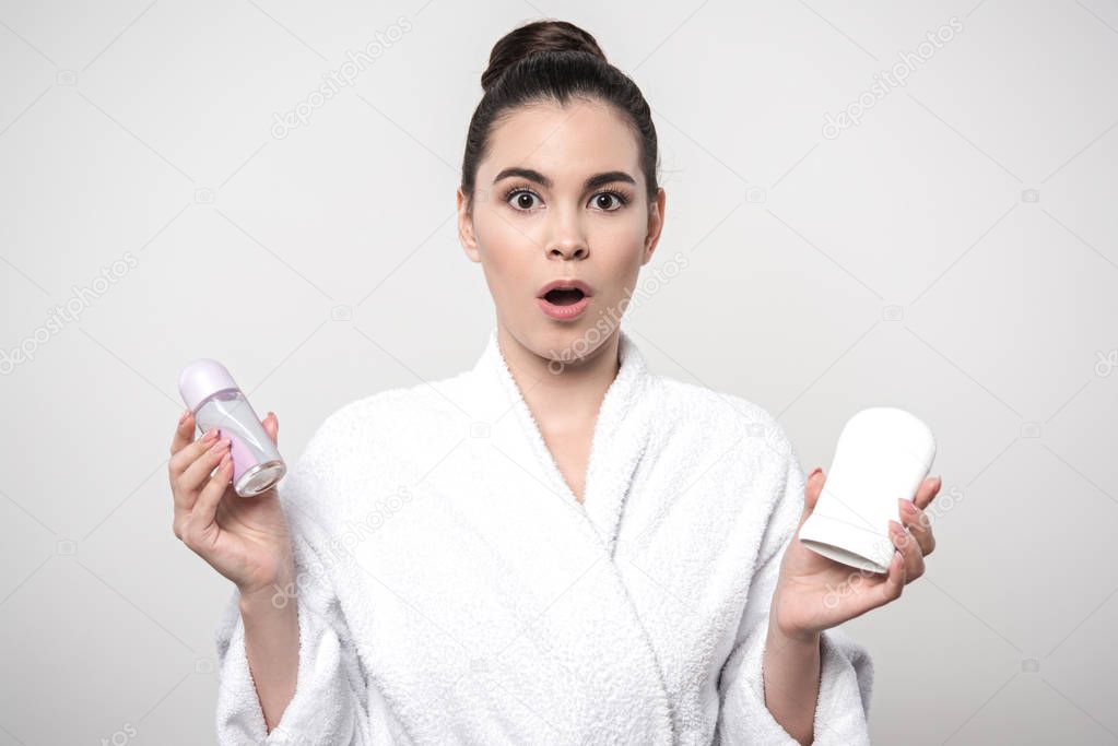 surprised woman in bathrobe holding deodorants while looking at camera isolated on grey
