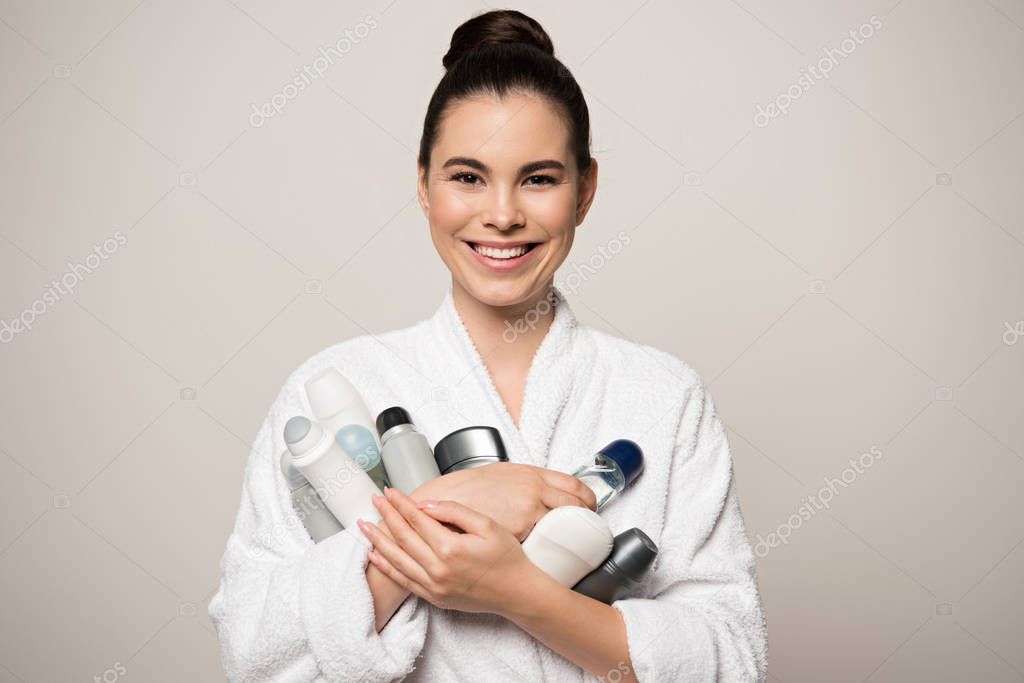 joyful woman in bathrobe holding different deodorants while looking at camera isolated on grey