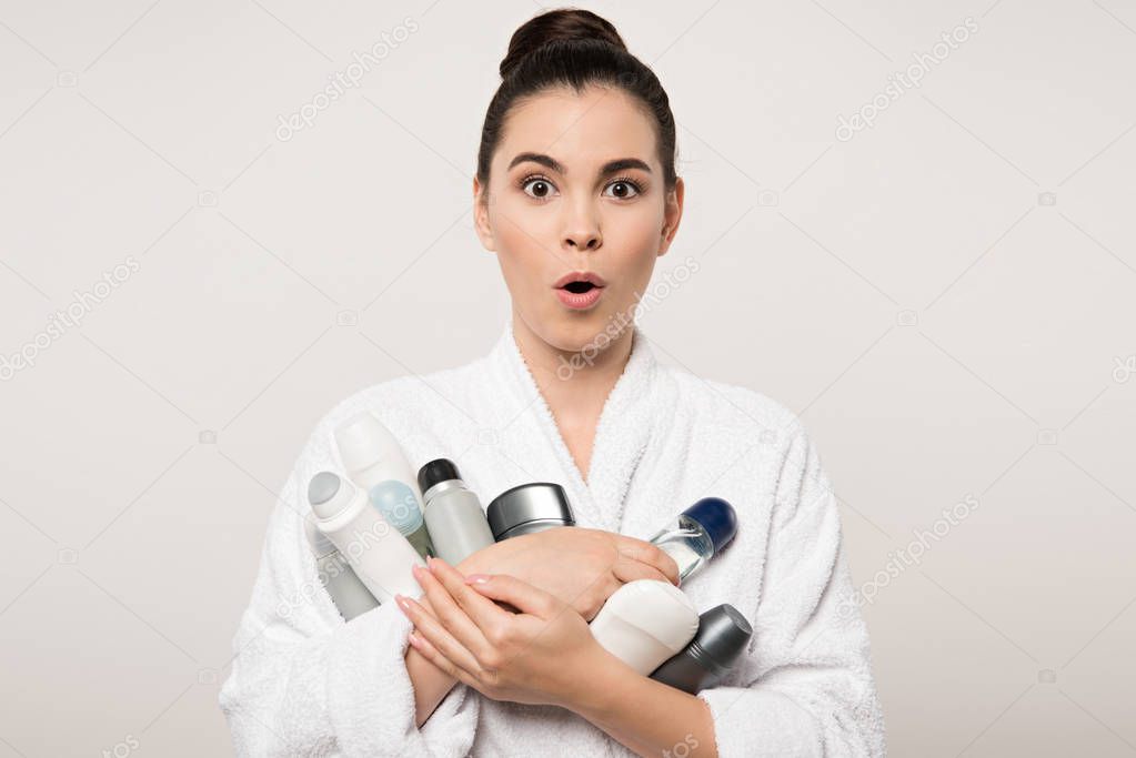 excited woman in bathrobe holding different deodorants while looking at camera isolated on grey