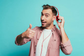 happy man listening music with headphones and showing thumb up, isolated on blue