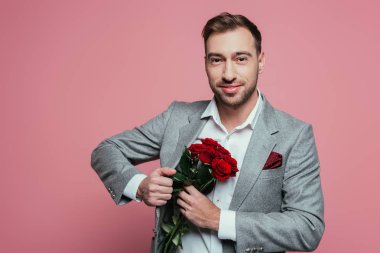 Kırmızı güller tutan takım elbiseli yakışıklı pozitif adam, pembe üzerinde izole edilmiş.