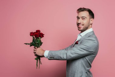 Kırmızı güller tutan takım elbiseli yakışıklı neşeli adam, pembe üzerinde izole edilmiş.