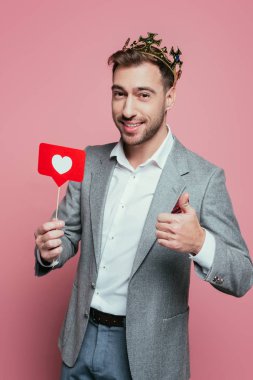 Taç giymiş mutlu bir adam baş parmağını gösteriyor ve sevgililer günü için kalpten kart tutuyor. Pembe renkte izole edilmiş. 