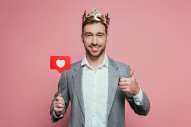 Taç giymiş mutlu bir adam baş parmağını gösteriyor ve sevgililer günü için kalpten kart tutuyor. Pembe renkte izole edilmiş. 