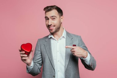 Sevgililer günü için kalp hediye kutusunu gösteren mutlu sakallı adam, pembe renkte izole edilmiş.