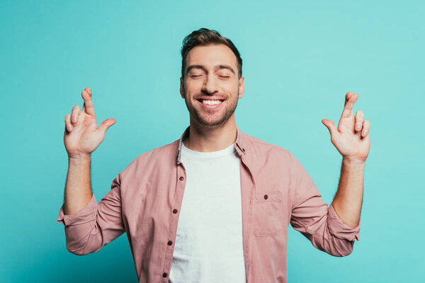 cheerful man with closed eyes and fingers crossed isolated on blue