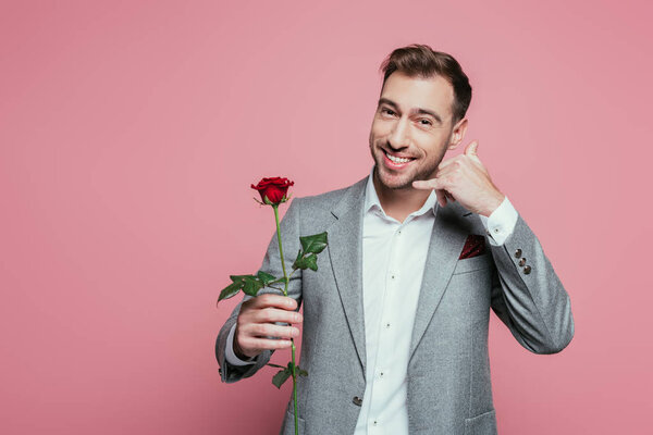 cheerful man holding rose and showing call me gesture, isolated on pink