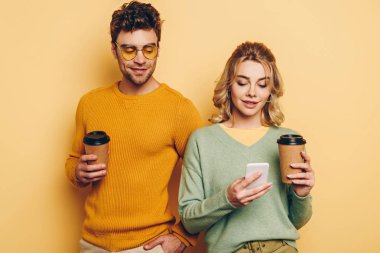 Gülümseyen bir çift kahve içerken, kız sarı arka planda akıllı telefondan sohbet ediyor.