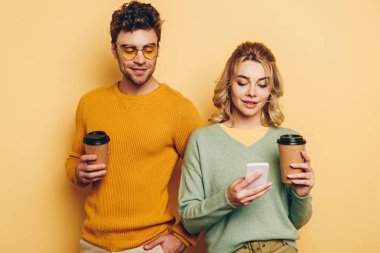 smiling couple holding coffee to go while girl chatting on smartphone on yellow background clipart