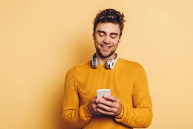 Boynunda kablosuz kulaklıkla gülümseyen adam sarı arka planda akıllı telefondan sohbet ediyor.