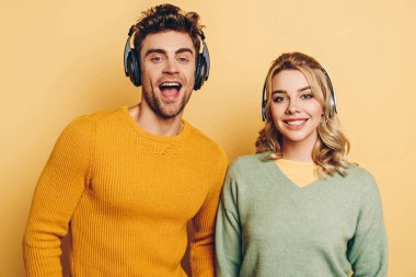 happy man and woman smiling at camera while listening music in wireless headphones on yellow background clipart