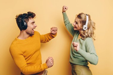 happy man and woman dancing while listening music in wireless headphones on yellow background clipart