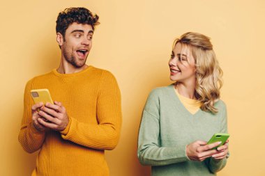 Gülümseyen adam ve kadın sarı arka planda akıllı telefonlardan sohbet ederken birbirlerine bakıyorlar.