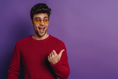 cheerful young man pointing with thumb and looking away on purple background clipart