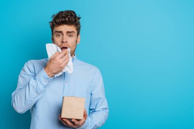 sick man sneezing in paper napkin while looking at camera on blue background clipart