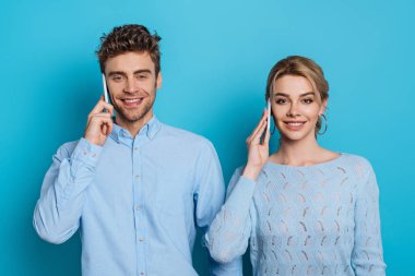 Genç adam ve kadın kameraya gülümserken mavi arka planda akıllı telefonlardan konuşuyorlar.