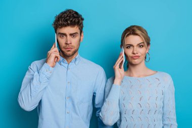 Kuşkucu adam ve kadın kameraya bakarken mavi arka planda akıllı telefonlardan konuşuyorlar.
