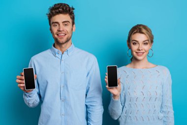 pozitif adam ve kadın mavi arkaplanda kameraya bakarken boş ekranlı akıllı telefonlar gösteriyor