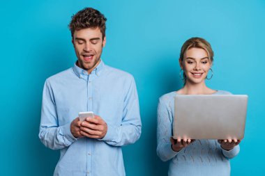 Neşeli bir adam mavi arka planda dizüstü bilgisayarı olan kız arkadaşının yanında akıllı telefondan sohbet ediyor.