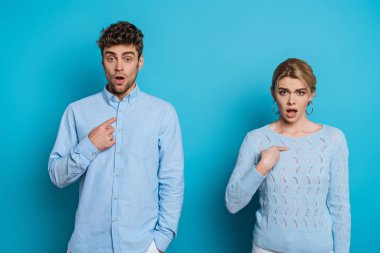 shocked man and woman pointing with fingers at themselves while looking at camera on blue background clipart