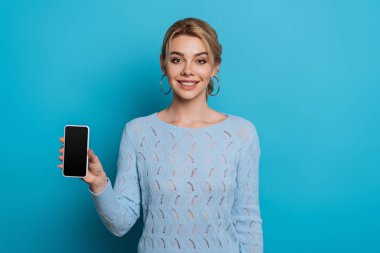 Mavi arka planda boş ekranlı akıllı telefonu gösterirken kameraya gülümseyen neşeli kız