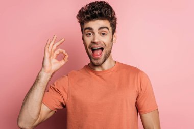 Neşeli genç adam pembe arka plandaki kameraya bakarken iyi bir jest yapıyor.