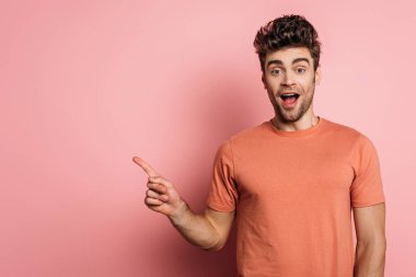 Heyecanlı genç adam pembe arka planı işaret ederken kameraya bakıyor.
