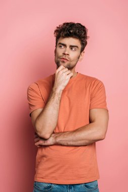 Düşünceli genç adam pembe arka plana bakarken çenesine dokunuyor.