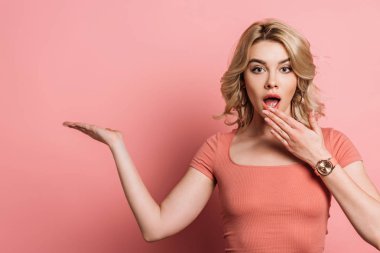 Şok olmuş bir kız, pembe arka planı işaret ederken eliyle ağzını kapatıyor.