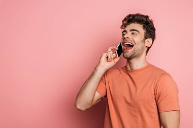 cheerful young man laughing while talking on smartphone on pink background clipart