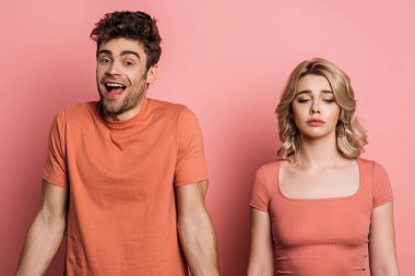 Neşeli adam pembe arka planda gücenmiş kız arkadaşının yanında dururken kameraya bakıyor.