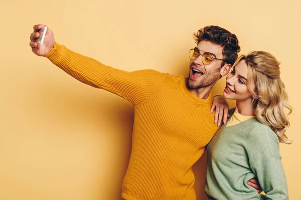 Neşeli adam akıllı telefondan selfie çekiyor ve sarı arka planda güzel bir kız arkadaşı var.