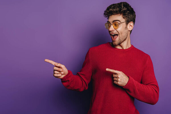 excited young man pointing with fingers and looking away on purple background