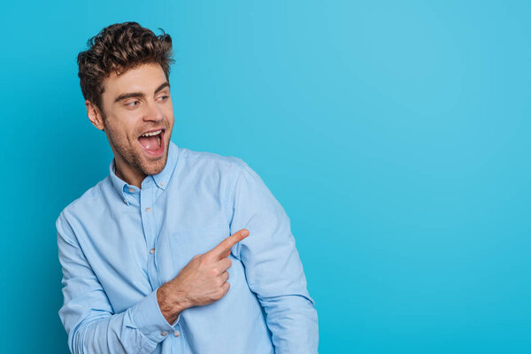 excited young man looking away and laughing while pointing with finger on blue background