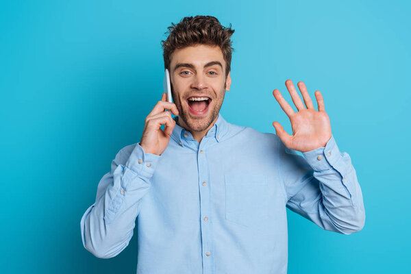 happy young man waving hand while talking on smartphone on blue background