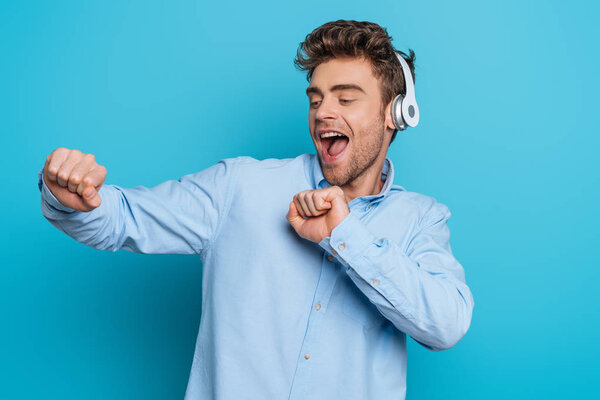 cheerful young man dancing in wireless headphones on blue background