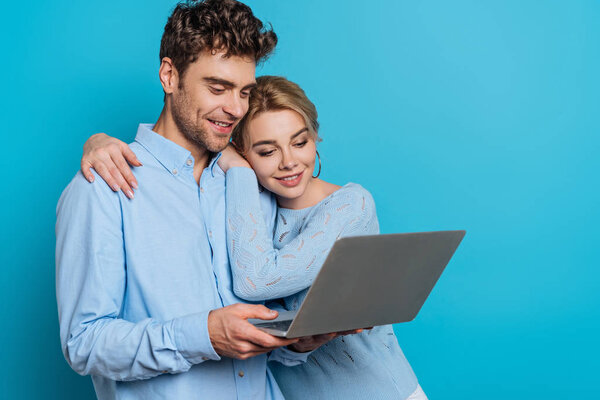 happy girl hugging smiling boyfriend while looking at laptop together on blue background