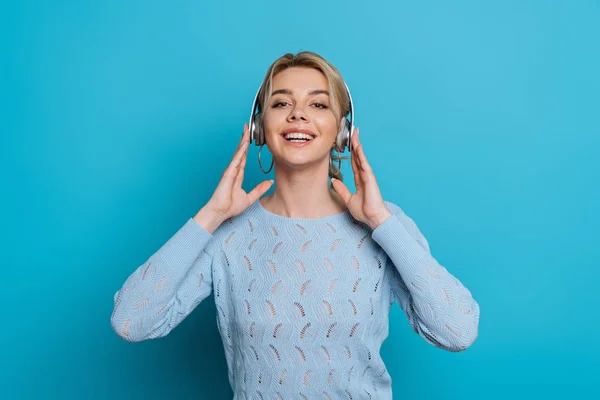 在蓝色背景的无线耳机里听音乐时 快乐的女孩对着相机微笑 — 图库照片