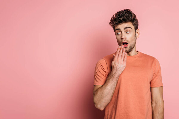 shocked young man holding hand near mouth while looking away on pink background