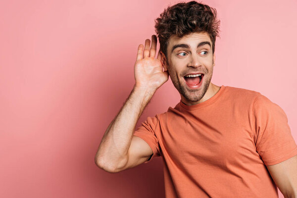 curious young man eavesdropping while holding hand near ear on pink background