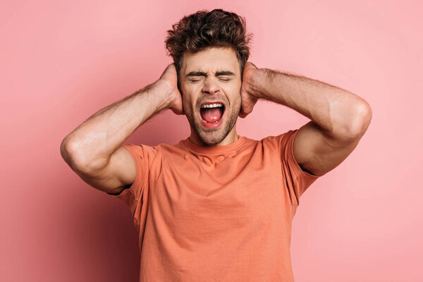 раздраженный молодой человек кричит и закрывает уши руками на розовом фоне
