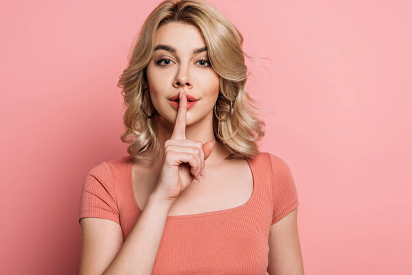 attractive girl looking at camera while showing hush gesture on pink background